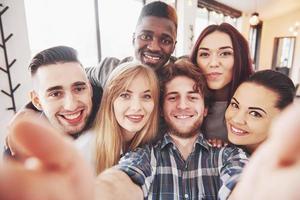 Friends having fun at restaurant.Two boys and four girls drinking making selfie, making peace sign and laughing. On foreground woman holding smart phone. All wear casual clothes photo