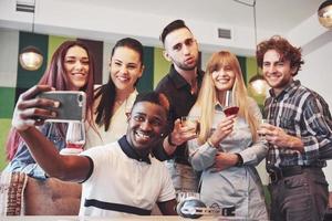 Friends having fun at restaurant.Two boys and four girls drinking making selfie, making peace sign and laughing. On foreground woman holding smart phone. All wear casual clothes
