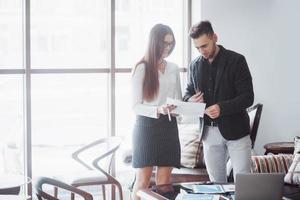 Businessman and business woman studying a chart on the plate and paper documents at the window on the background of the city office on a high floor photo