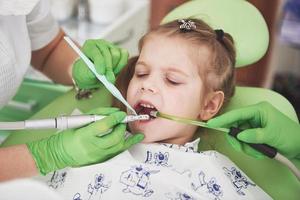 Manos de dentista pediátrico irreconocible y asistente que hace el procedimiento de examen para una niña linda sonriente sentada en una silla en el hospital foto