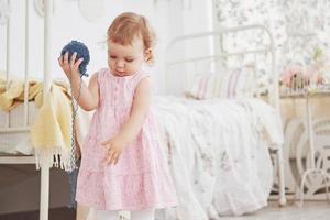 Childhood concept. Baby girl in cute dress play with colored thread. White vintage childroom photo