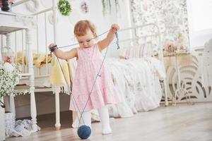Childhood concept. Baby girl in cute dress play with colored thread. White vintage childroom