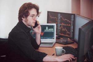 Over the shoulder view of and stock broker trading online while accepting orders by phone. Multiple computer screens ful of charts and data analyses in background photo