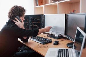 Over the shoulder view of and stock broker trading online while accepting orders by phone. Multiple computer screens ful of charts and data analyses in background photo
