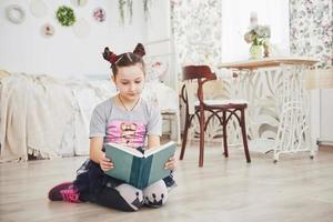 Cute little child girl reading a book in the bedroom. Kid with crown sitting on the bed near window photo