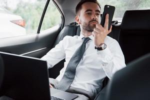 A young businessman working on laptop and talking on the phone while sitting in the car's back. Works in motion, appreciates its time photo