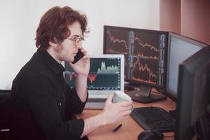 Over the shoulder view of and stock broker trading online while accepting orders by phone. Multiple computer screens ful of charts and data analyses in background photo