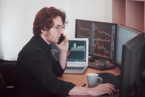 Over the shoulder view of and stock broker trading online while accepting orders by phone. Multiple computer screens ful of charts and data analyses in background photo