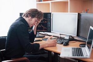 Stressful day at the office. Young businessman holding hands on his face while sitting at the desk in creative office. Stock Exchange Trading Forex Finance Graphic Concept photo