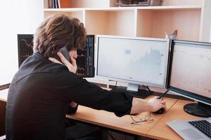 Over the shoulder view of and stock broker trading online while accepting orders by phone. Multiple computer screens ful of charts and data analyses in background photo