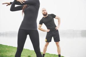 Fitness couple stretching outdoors in park near the water. Young bearded man and woman exercising together in morning photo