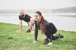 Fitness couple stretching outdoors in park near the water. Young bearded man and woman exercising together in morning photo