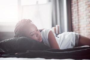 A beautiful African woman lies and sleeps in bed, in a white bedroom. Good morning, vacation at home. The girl is wearing a T-shirt, pigtails on her head photo