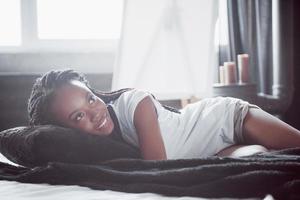 una hermosa mujer africana yace y duerme en la cama, en un dormitorio blanco. buenos dias vacaciones en casa. la niña lleva una camiseta, coletas en la cabeza