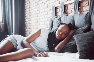 una hermosa mujer africana yace y duerme en la cama, en un dormitorio blanco. buenos dias vacaciones en casa. la niña lleva una camiseta, coletas en la cabeza