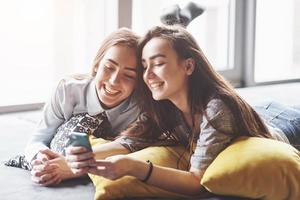 dos lindas hermanas gemelas sonrientes sosteniendo smartphone y haciendo selfie. las niñas se acuestan en el sofá posando y alegría foto