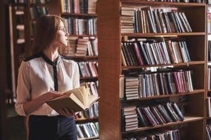 Joven y atractiva bibliotecaria estudiante leyendo un libro entre las estanterías de la biblioteca foto