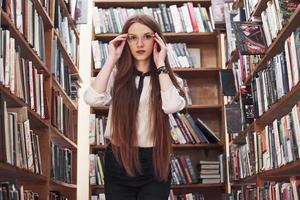 Joven y atractiva bibliotecaria estudiante leyendo un libro entre las estanterías de la biblioteca foto