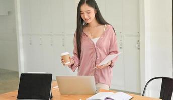 una estudiante sosteniendo una taza de café y un documento de mano con una computadora portátil sobre la mesa.