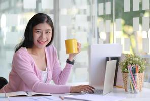 una joven ejecutiva sostiene una taza de café y sonríe mientras usa una computadora portátil en su oficina personal. foto