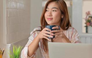 estudiante adolescente sonríe y bebe café mientras estudia en línea desde su computadora portátil en casa. foto