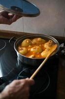 Aged woman's hands with a cooking pot and a wooden spoon while cooking potatoes filled with tuna, tomato and eggs. Gastronomy at home. Typical asturian dish photo