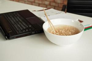 Los fideos instantáneos en un tazón y los palillos se colocan cerca de la computadora portátil en el escritorio de trabajo blanco en la oficina para una hora de almuerzo apresurada y desordenada porque se cocina rápidamente y es una comida sabrosa y de estilo de vida poco saludable. foto