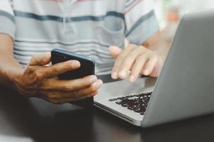 Mano de hombre usando un teléfono inteligente y una computadora portátil en la mesa en casa, buscando información navegando por Internet en la web, trabajando desde casa concepto de compras en línea de negocios foto