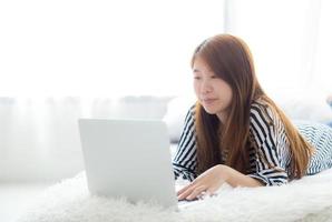 joven asiática acostada en la cama usando la computadora portátil en el dormitorio. foto