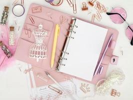 Top view of a pink planner with cute stationery. Pink glamour planner with a white mannequin figurine. Planner with open pages on a white background and with beautiful accessories pens, buttons, pins. photo