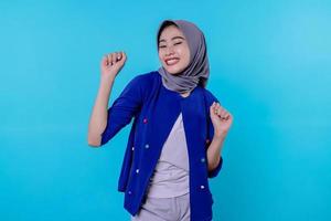 Cheerful young female has positive expression, clenches fists, has overjoyed look, being in high spirit, wearing hijab, isolated over blue wall photo