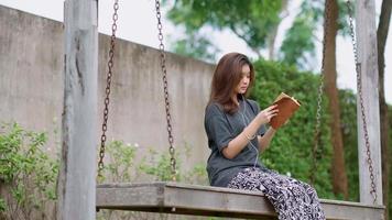 uma adolescente asiática lê um livro enquanto está sentada em um balanço em seu jardim.