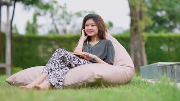 una hermosa mujer asiática disfruta escuchando música con auriculares y se siente feliz y relajada al aire libre en el jardín de su casa. video