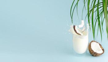 salpicaduras de leche, trozos de coco volando sobre el vaso sobre fondo azul de verano con hojas de palma. copie el espacio.