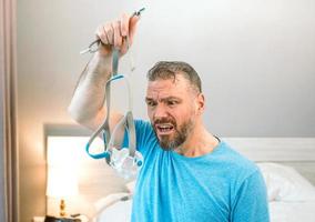 Unhappy shocked man with chronic breathing issues surprised by using  CPAP machine sitting on the bed in bedroom. Healthcare, CPAP, Obstructive sleep apnea therapy, snoring concept photo