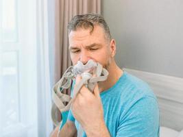 Unhappy shocked man with chronic breathing issues surprised by using  CPAP machine sitting on the bed in bedroom. Healthcare, CPAP, Obstructive sleep apnea therapy, snoring concept photo