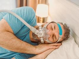 Sleeping man with chronic breathing issues considers using CPAP machine in bed. Healthcare, Obstructive sleep apnea therapy, CPAP, snoring concept photo