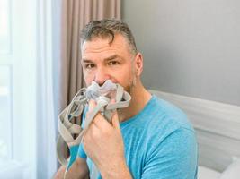 Unhappy shocked man with chronic breathing issues surprised by using  CPAP machine sitting on the bed in bedroom. Healthcare, CPAP, Obstructive sleep apnea therapy, snoring concept photo