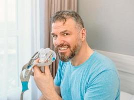 Mature happy man with chronic breathing issues considers using sitting on the bed in bedroom with CPAP machine. Healthcare, CPAP, snoring concept photo