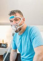 Unhappy shocked man with chronic breathing issues surprised by using  CPAP machine sitting on the bed in bedroom. Healthcare, CPAP, Obstructive sleep apnea therapy, snoring concept photo