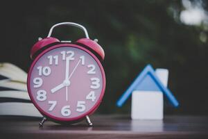 reloj despertador y modelo de casa y libros sobre la mesa de madera con fondo bokeh. foto