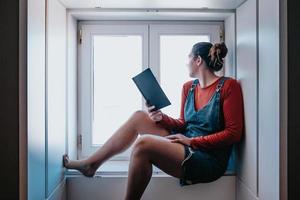 Mujer joven sentada junto a una ventana leyendo un libro durante un día brillante, conceptos de reflexión y autocuidado, espacio de copia foto