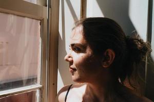 Young woman looking through a window during a super sunny day, copy space, reflexion and thinking concept, sad and anxiety, self care photo