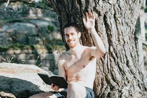 A young hippie man in trunks shirtless reading a book against a tree and saluting to camera during a summer day, relax and chill concepts, good life, young people