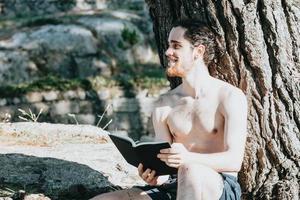A man in trunks shirtless reading a book against a tree during a summer day, relax and chill concepts, good life, young people, looking away from camera copy space