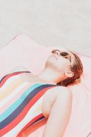 imagen de arriba de una mujer joven en un colorido traje de baño tomando un baño de sol mientras usa el concepto de gafas de sol, vacaciones y descanso. marketing en redes sociales, viajes modernos de bajo costo foto