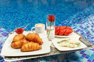 croissants, mermelada, queso, fresa y café de pie sobre una bandeja al borde de una piscina. foto