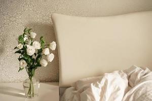 white flowers in a transparent glass vase stand on a bedside table by a bed with a large white headboard. bedroom interior. modern room photo