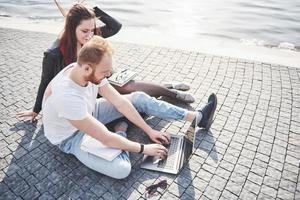 Two students guy and girl are sitting outdoors and enjoying a laptop, studying outdoors on a sunny day photo