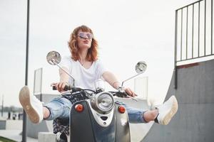 Retrato de una hermosa chica hipster sentada en un scooter retro negro, sonriendo posando y disfrutando del cálido sol primaveral.
